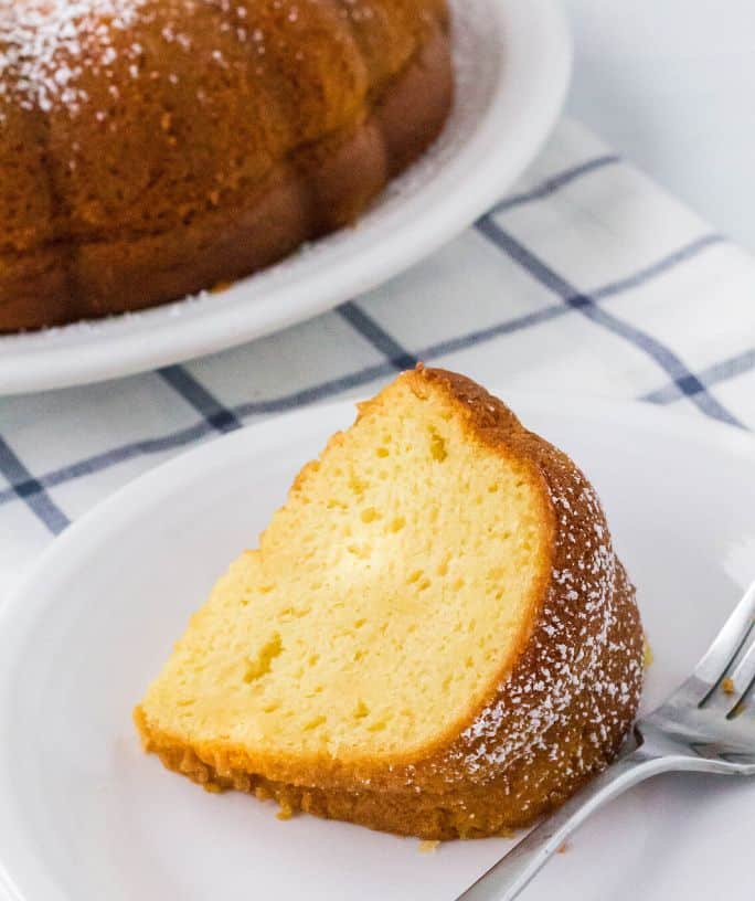 slice of vanilla pudding bundt cake on a white plate, with the remainder of the bundt cake in the background