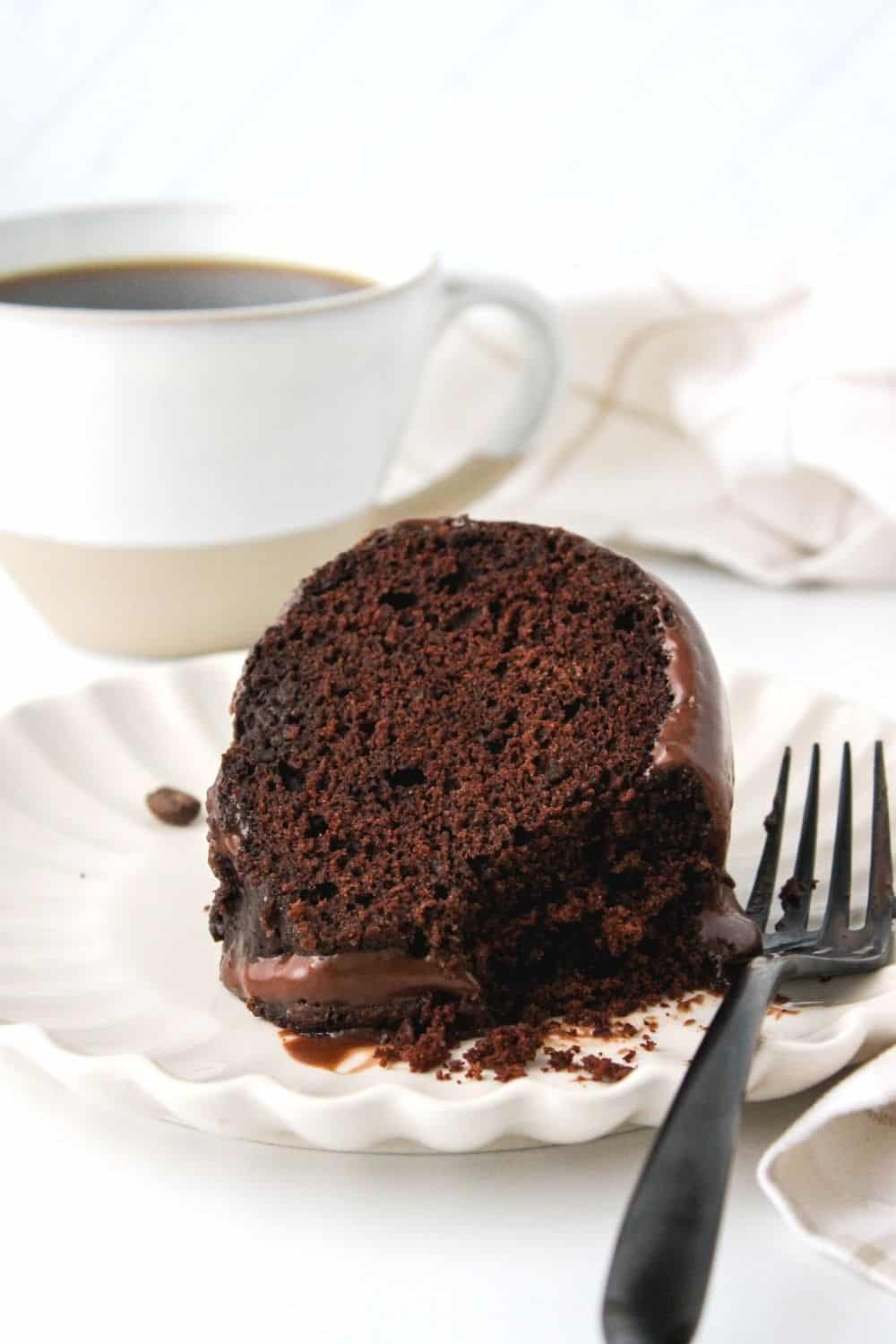 slice of chocolate fudge cake on a white plate, with a cup of coffee in the background