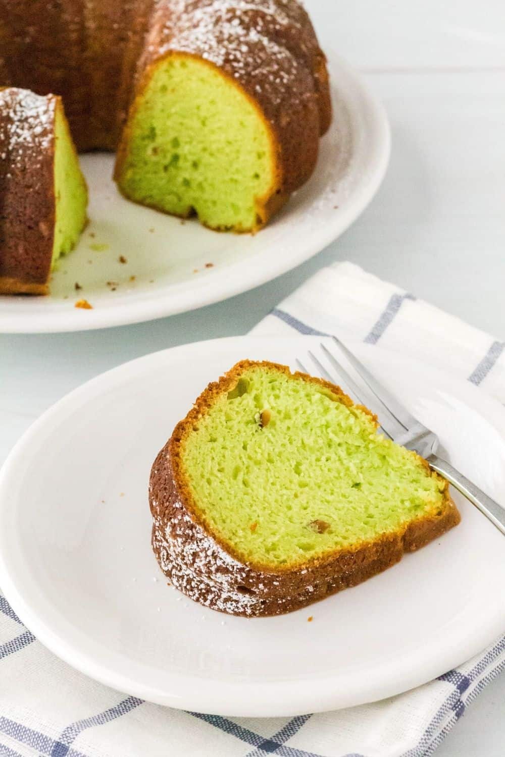 slice of pistachio bundt cake on a white plate with a fork, and the remainder of the cake in the background