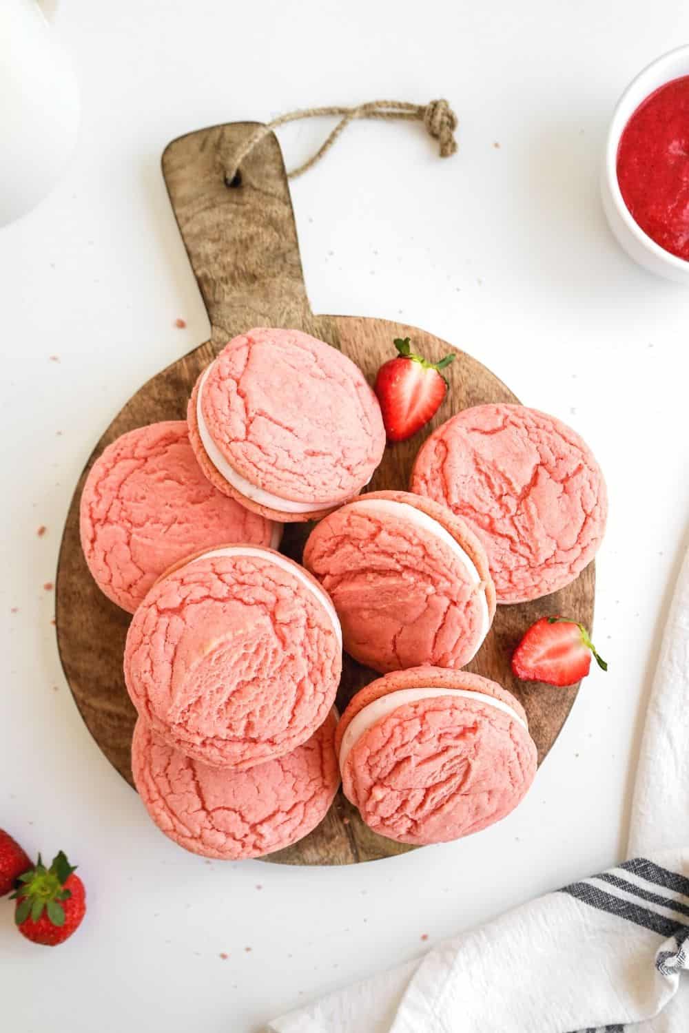 seven strawberry sandwich cookies made with cake mix displayed on a wooden serving platter, with pieces of fresh strawberries scattered around