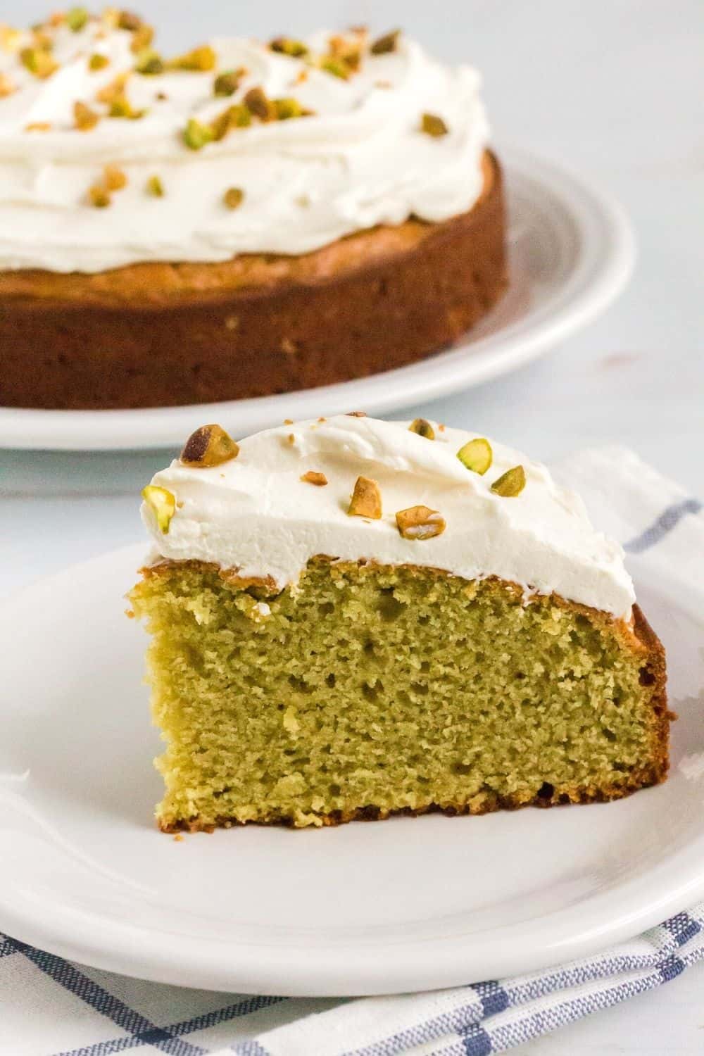 slice of pistachio cake made from scratch and topped with frosting, served on a white plate with the remainder of the single layer cake in the background