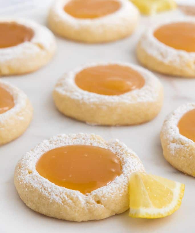 close-up view of several lemon curd cookies on a marble slab. A piece of fresh lemon rests beside one cookie.