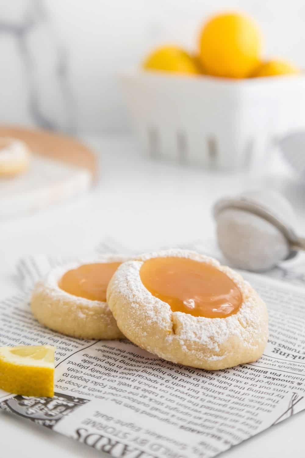 two lemon curd cookies resting on a piece of newsprint, with a powdered sugar duster and fresh lemons in the background 