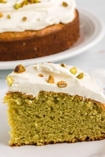slice of italian pistachio cake topped with mascarpone frosting; served on a white plate with whole cake in the background