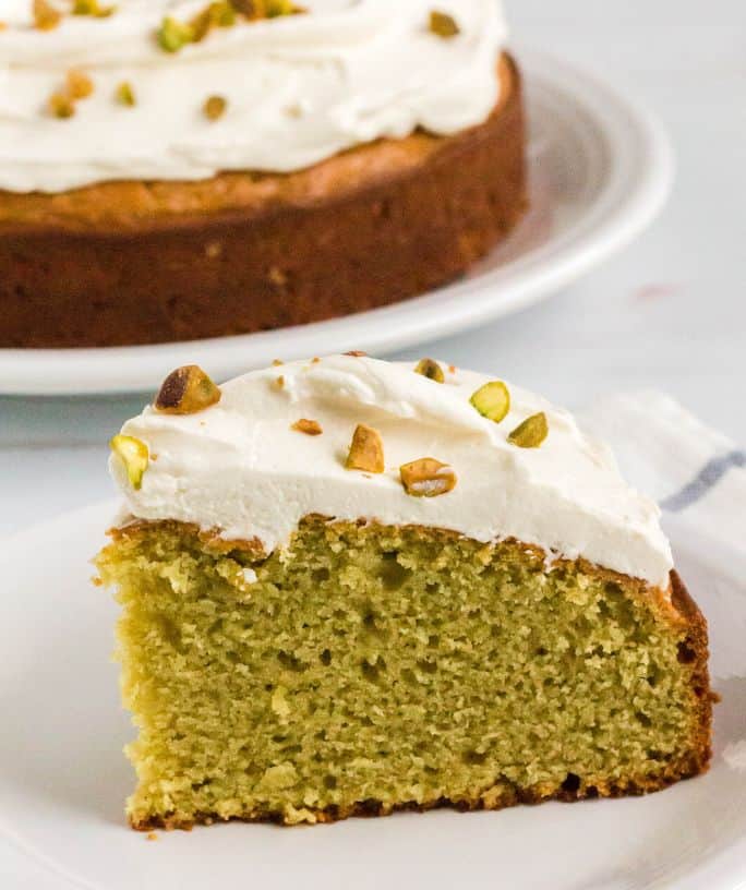 slice of italian pistachio cake topped with mascarpone frosting; served on a white plate with whole cake in the background