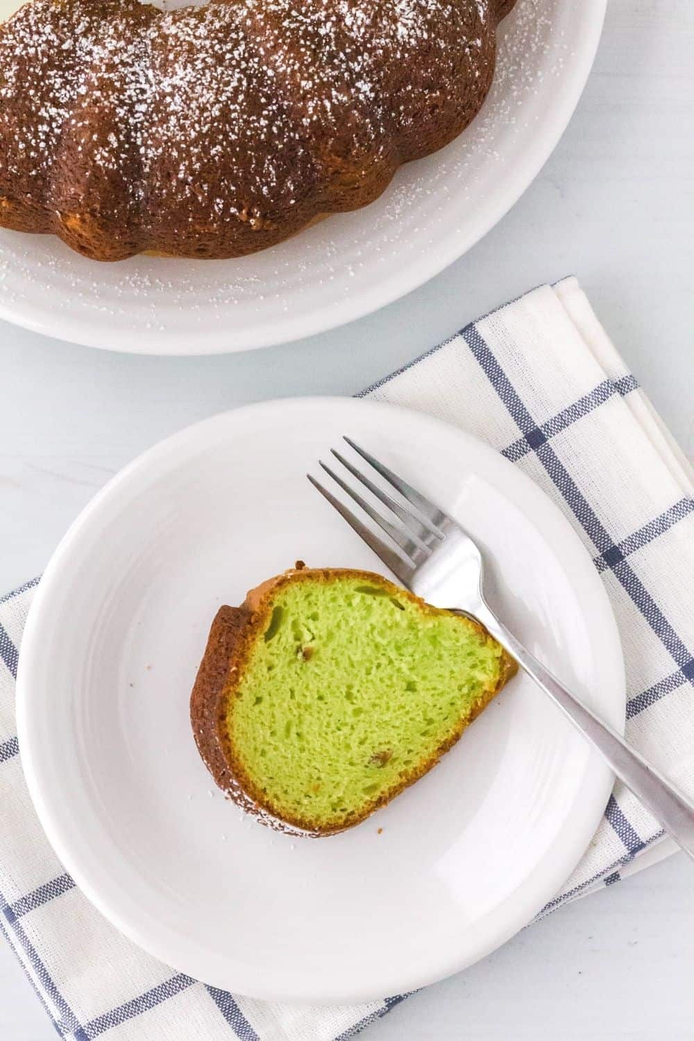overhead view of a slice of pistachio cake made from a mix, served on a white plate with a fork. The plate is resting on a blue and white napkin, and the remainder of the whole cake is in the background.