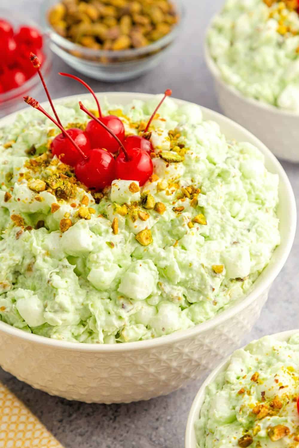 close-up view of a white bowl filled with watergate pistachio fluff salad, a pastel green creamy fruit salad topped with cherries and nuts.