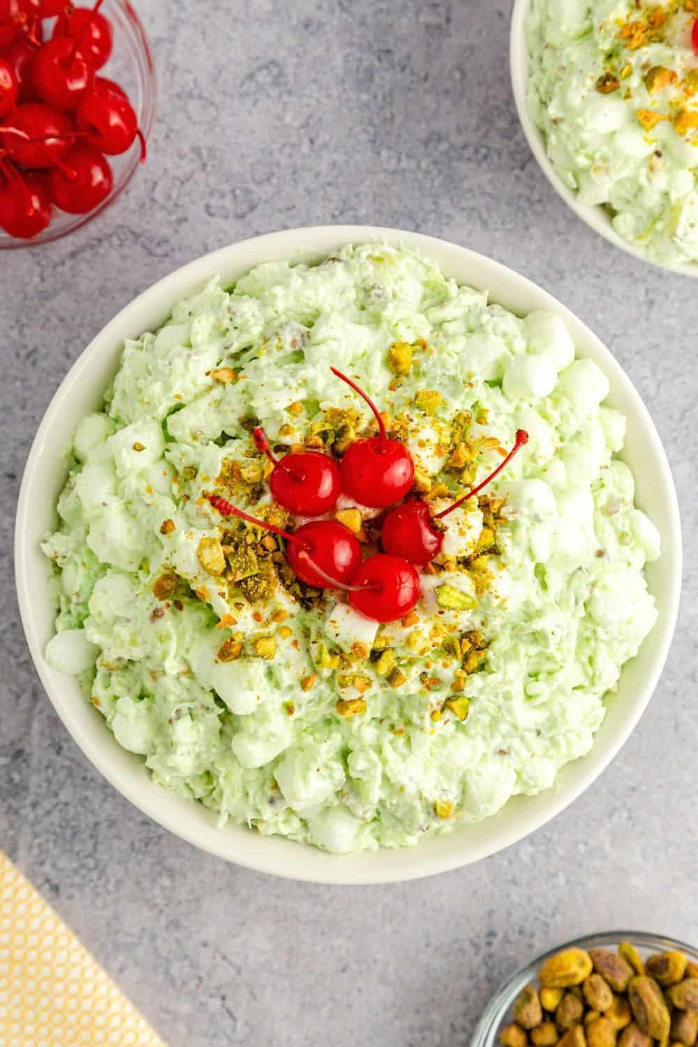 overhead view of a white bowl of watergate salad made with pineapple and marshmallows, garnished with cherries and chopped nuts