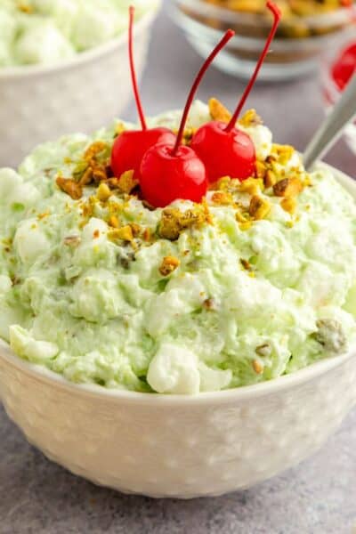 white bowl serving pistachio fluff salad, also known as watergate salad. It's made with pistachio pudding, marshmallows, whipped topping, pineapple, and topped with cherries and chopped pistachios.