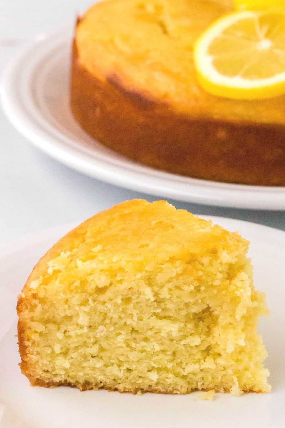 close-up view of a slice of lemon drizzle cake on a white plate, with the remaining syrup cake in the background.