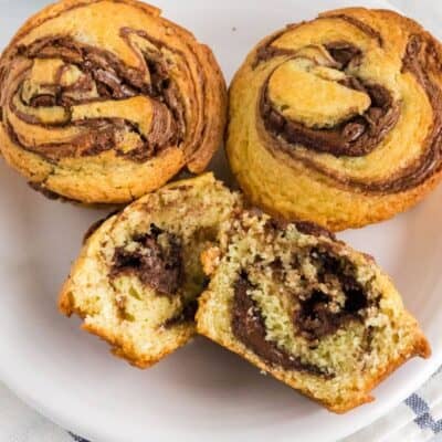 three Nutella muffins on a white plate; one muffin is cut in half to reveal the chocolate hazelnut swirl inside.