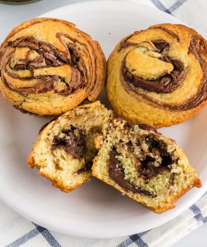 three Nutella muffins on a white plate; one muffin is cut in half to reveal the chocolate hazelnut swirl inside.