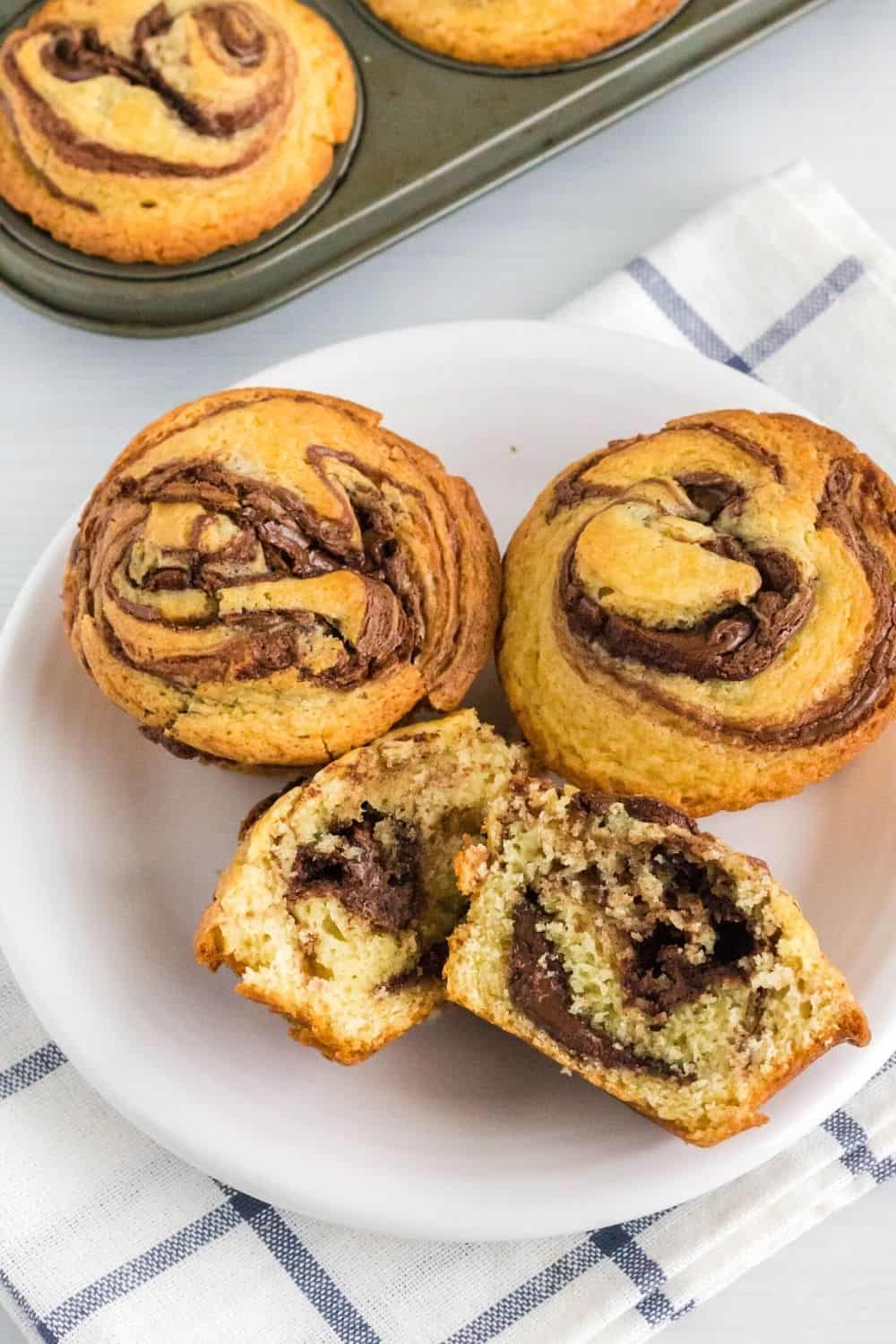 three Nutella stuffed muffins on a plate, with one muffin cut in half to reveal the Nutella swirl inside the muffin