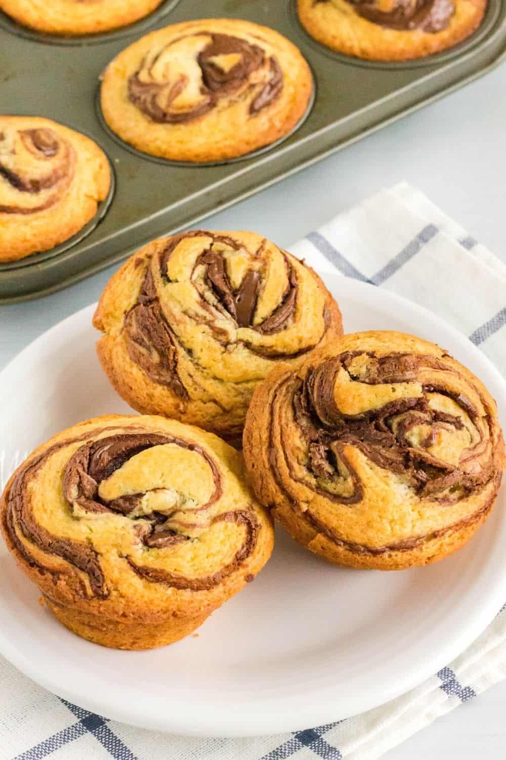 three swirled Nutella muffins are served on a white plate, with the muffin pan of remaining muffins in the background.
