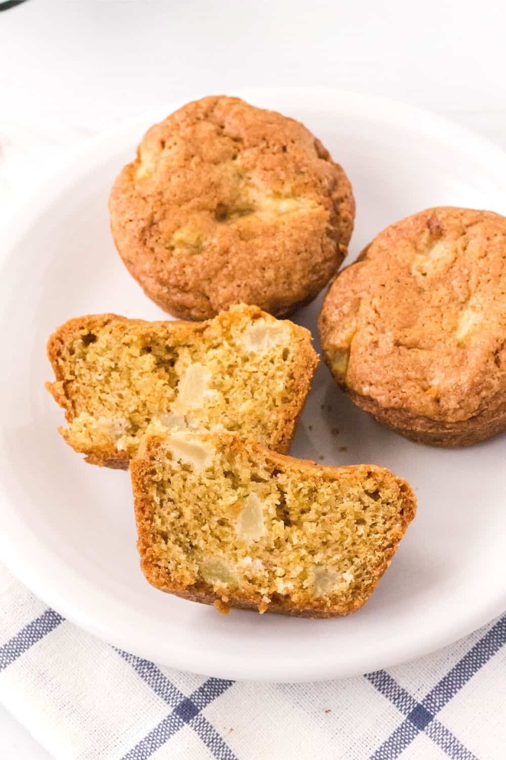 Pear muffins without streusel served on a white plate. One muffin is cut in half, showing the tender interior dotted with diced pears.