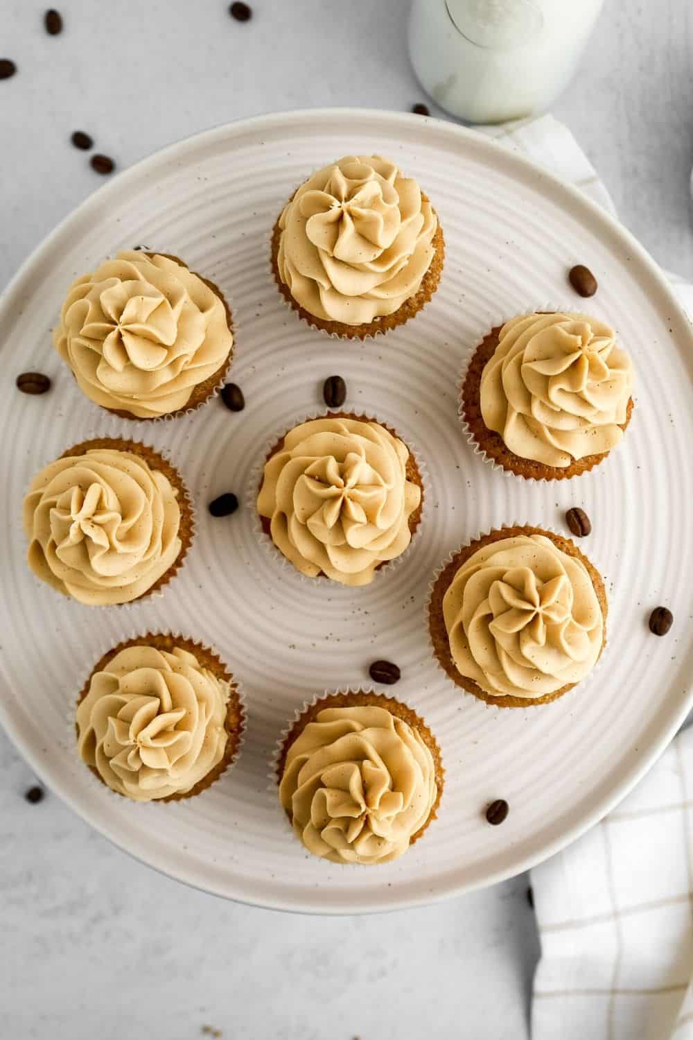 overhead view of several homemade coffee cupcakes on a cake plate, with coffee beans scattered around