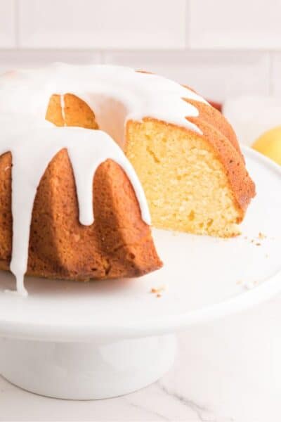 lemon sour cream pound cake on a white cake stand, drizzled with glaze. A slice has been cut out of the cake.