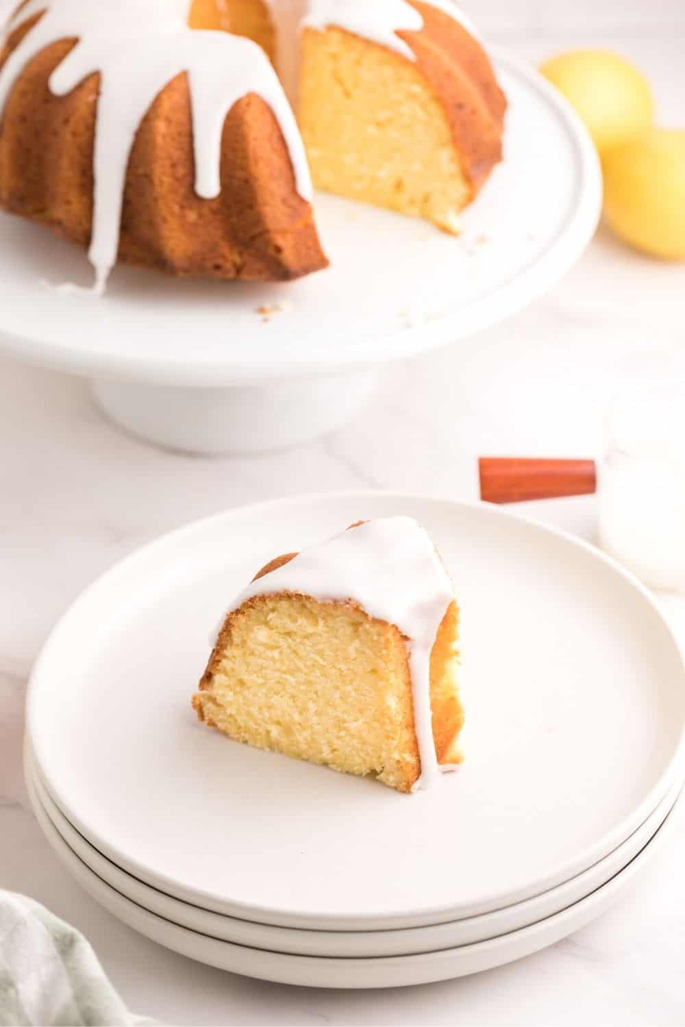 a slice of glazed lemon sponge cake served on a white plate, with the remaining sponge cake in the background.