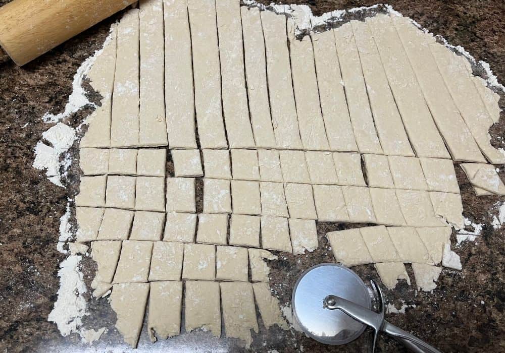 dumpling dough rolled out thin on a floured surface, and a pizza cutter cuts squares of dumplings