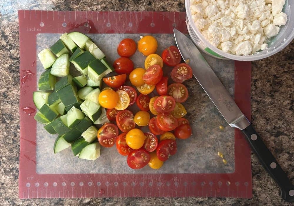 chopped cucumbers and tomatoes on a cutting board, with a container of feta crumbles next to it