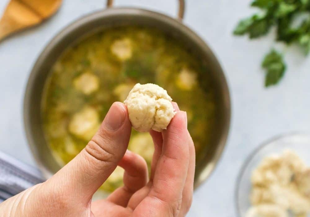a woman's hand holds a ball of dumpling dough over simmering broth