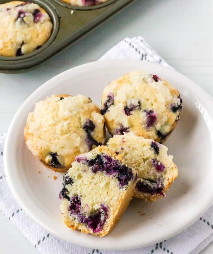 blueberry bisquick muffins on a white plate, with one muffin cut in half to show the interior.