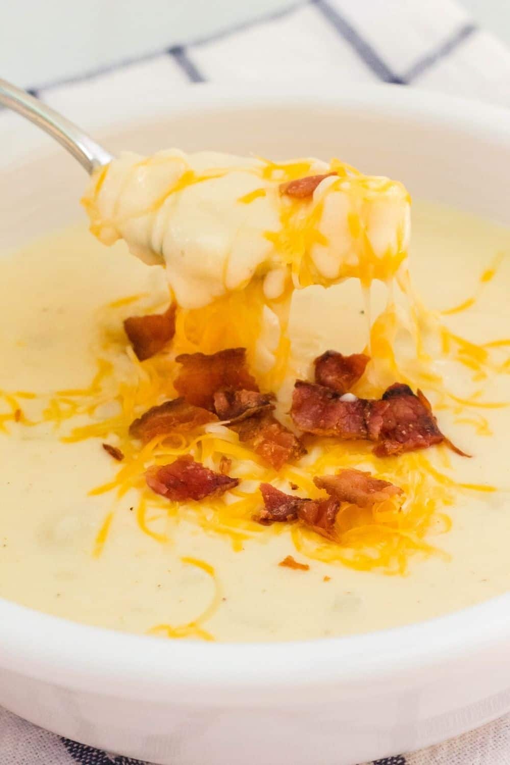 a spoon lifts a bite of homemade potato soup out of a white bowl. It's garnished with bacon and cheese, and is made from frozen hashbrowns.