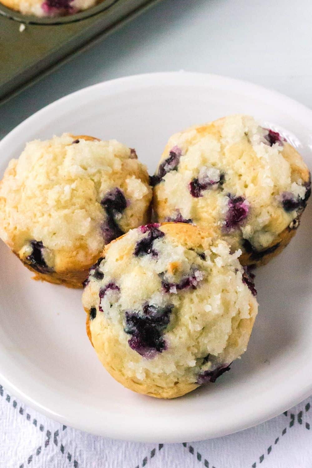 three whole bisquick blueberry muffins on a white plate, showing the sugar streusel topping.