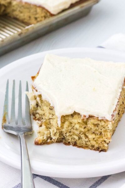 a slice of banana sheet cake served on a white plate with a fork. A bite has been taken out of the piece of cake.
