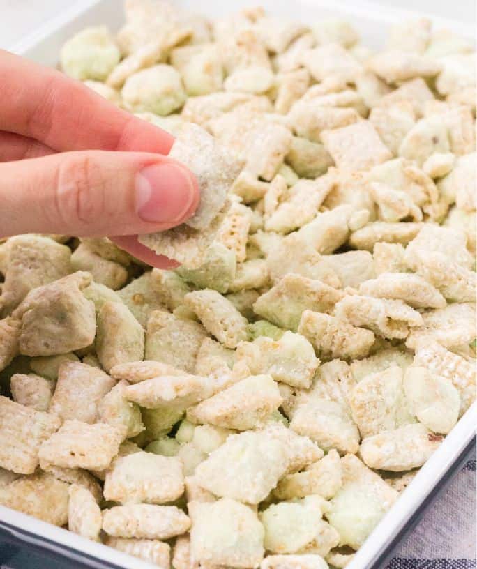 a woman's fingers lift out pistachio puppy chow pieces from a sheet pan serving the snack mix.