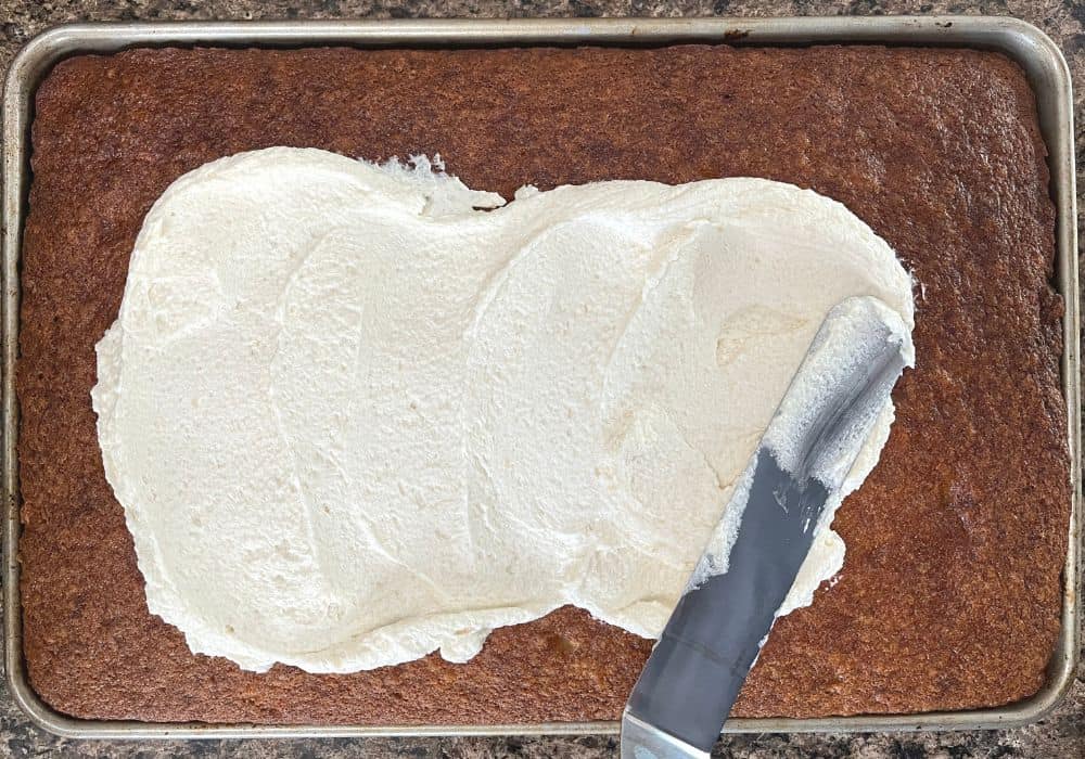 an offset spatula spreads the boiled milk frosting over the surface of the banana sheet cake.