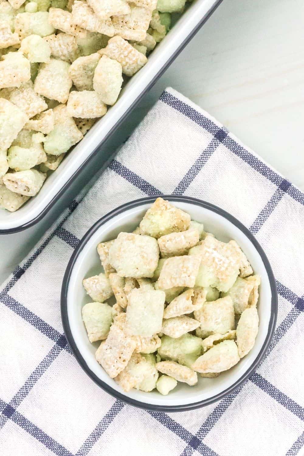 overhead view of a small dish serving pistachio puppy chow, with a sheet pan of remaining snack mix in the background.