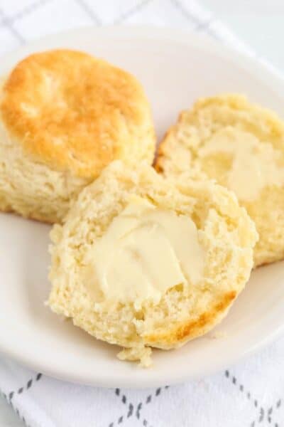 two Bisquick biscuits on a white plate; one is cut in half and spread with butter.