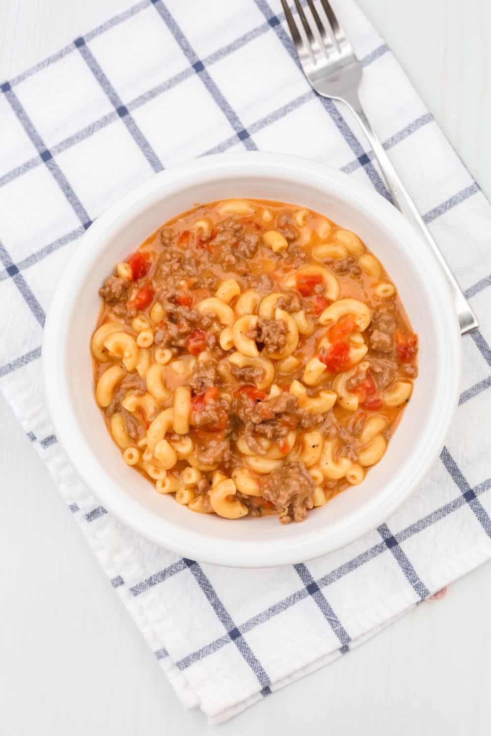 a white bowl of pressure cooker cheeseburger macaroni on top of a blue and white checkered napkin, with a fork next to it.