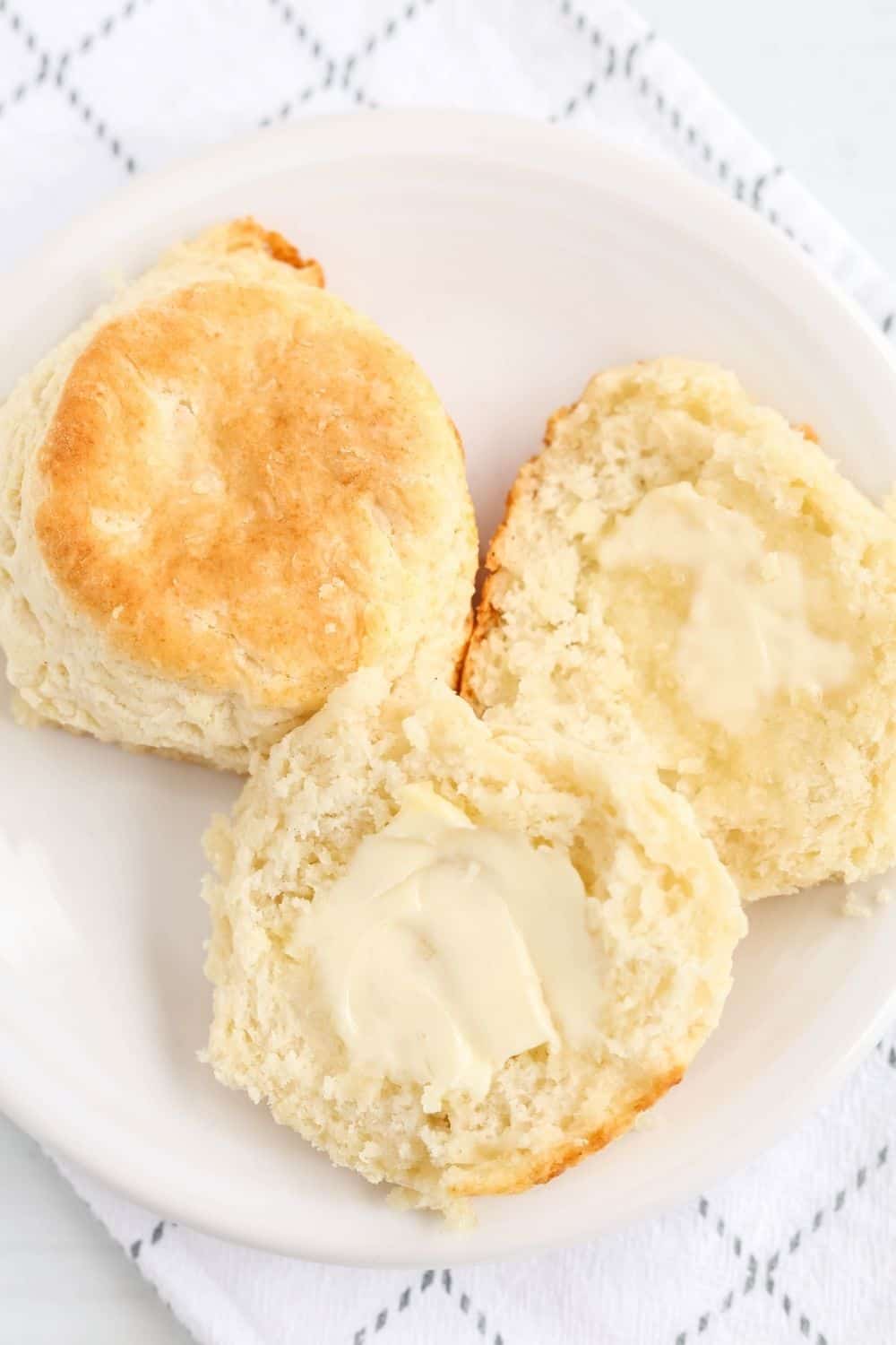 two bisquick biscuits on a white plate. One is cut in half, showing the fluffy, tender interior, spread with butter.