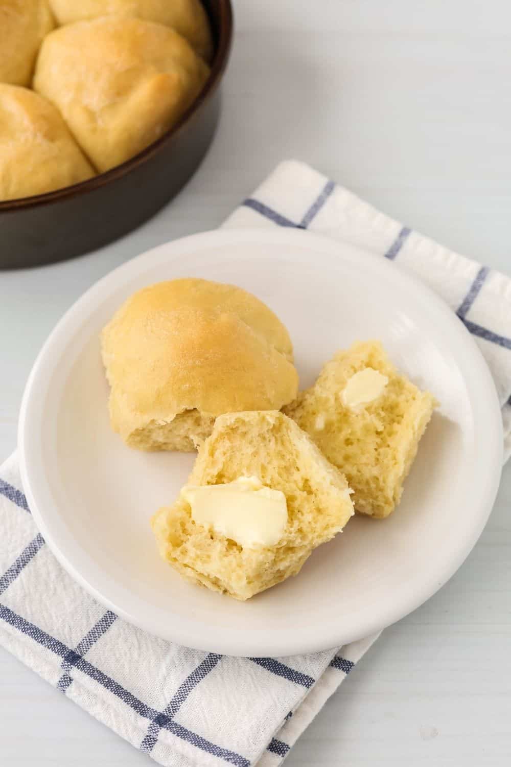 two homemade dinner rolls (that were previously proofed in the instant pot) served on a white plate, with the remaining pan of rolls in the background.