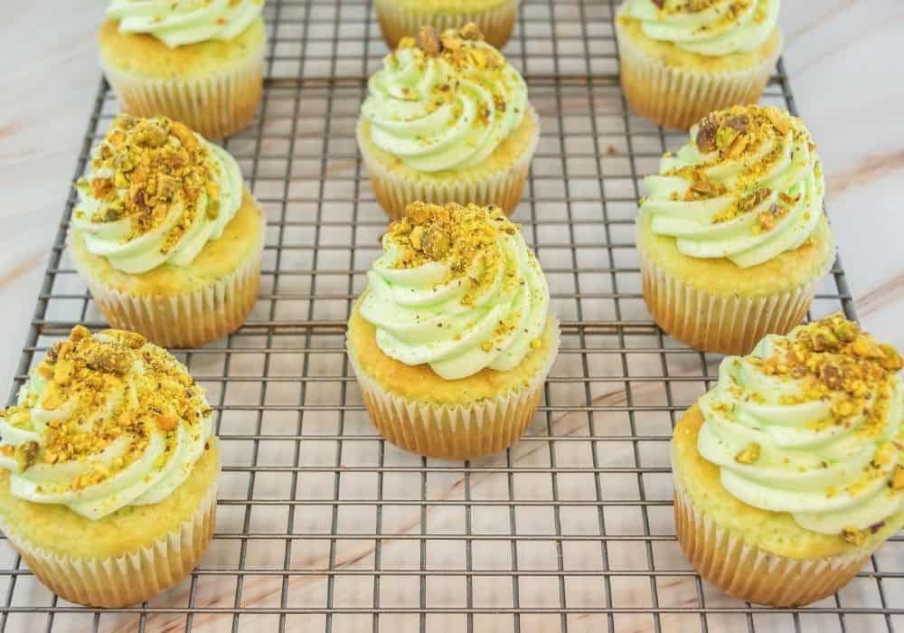 pistachio cupcakes topped with pistachio buttercream and chopped pistachios, displayed on a wire rack.