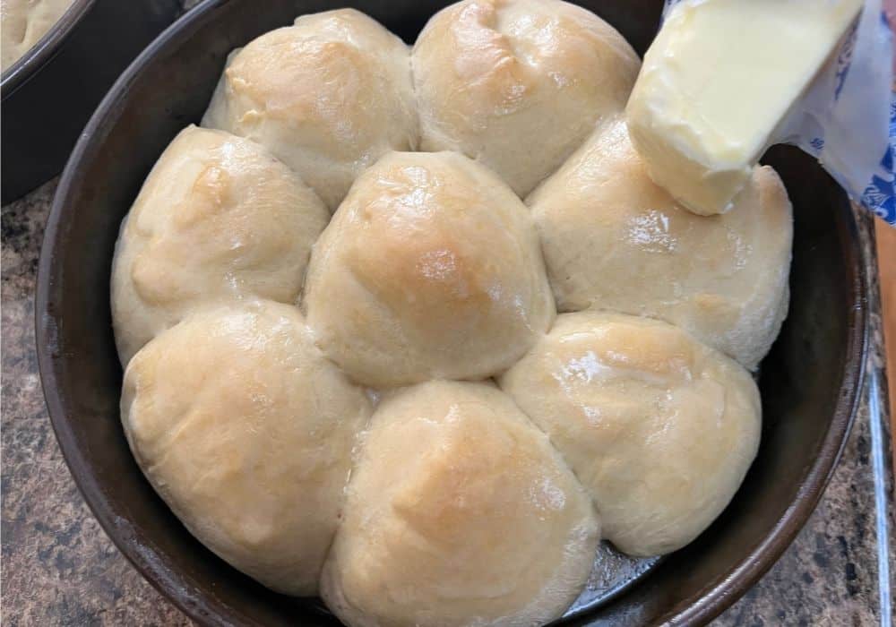 a stick of butter being rubbed over the tops of warm dinner rolls