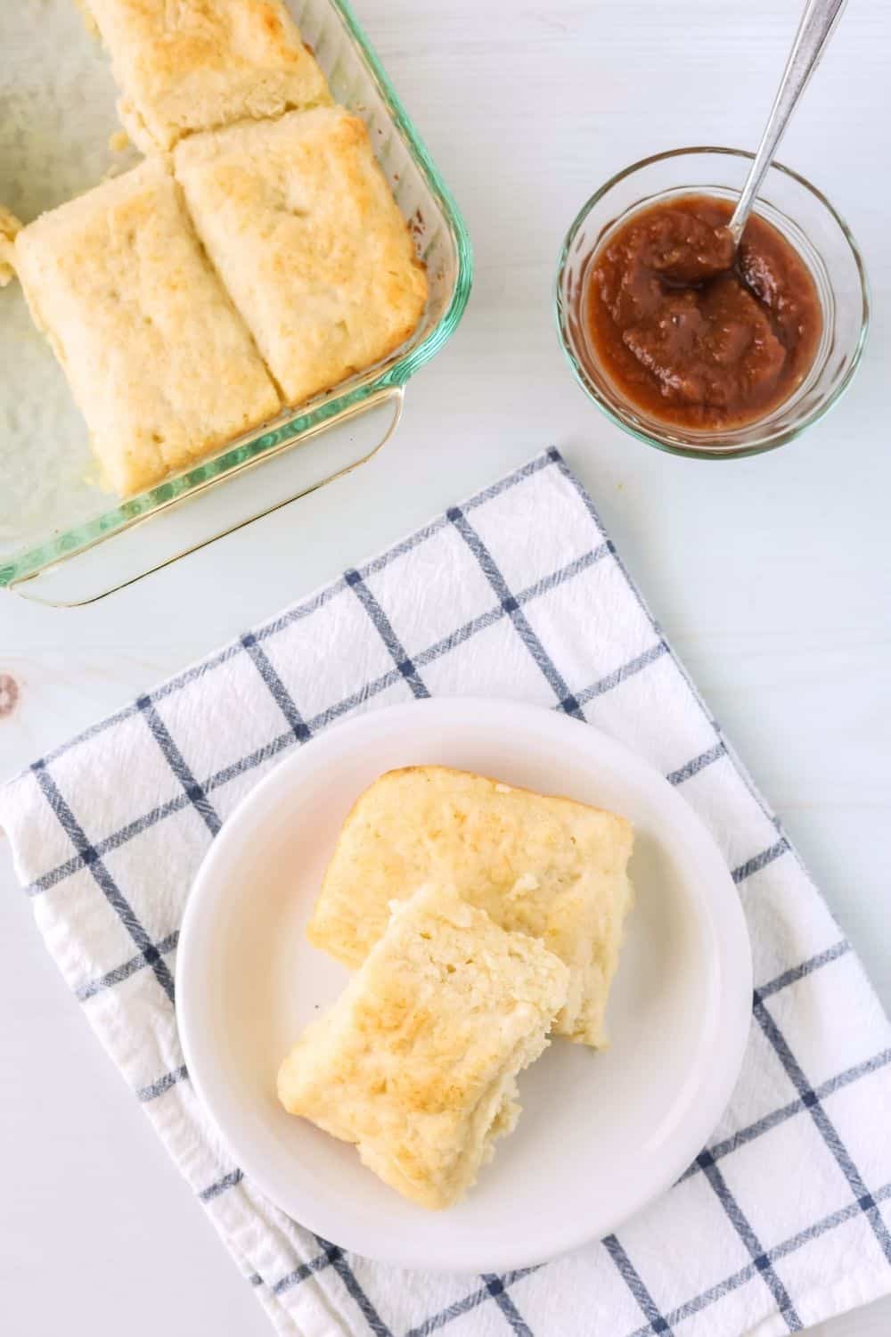 two fluffy 7Up and Bisquick biscuits on a white plate, with the baking dish of other biscuits in the background, along with a dish of apple butter.