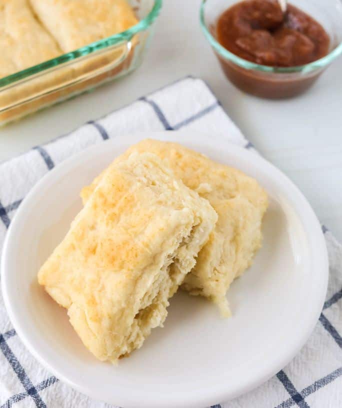 two 7Up biscuits on a white plate, with the pan of biscuits and a small dish of apple butter in the background.