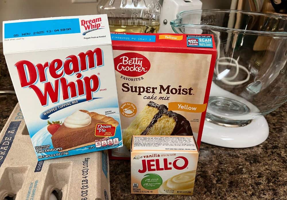 some of the ingredients needed for Dream Whip cake, including Dream Whip mix, cake mix, pudding mix, eggs, and oil. A stand mixer is in the background.