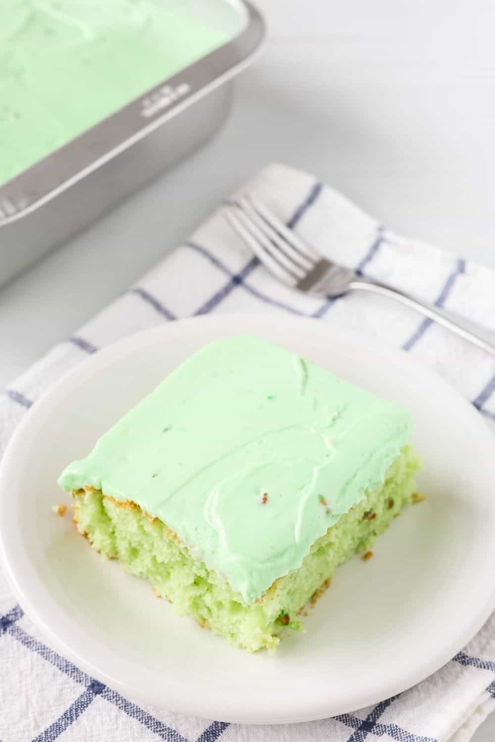 slice of pistachio pudding cake with pistachio frosting, served on a white plate. The remaining pan of cake is in the background.