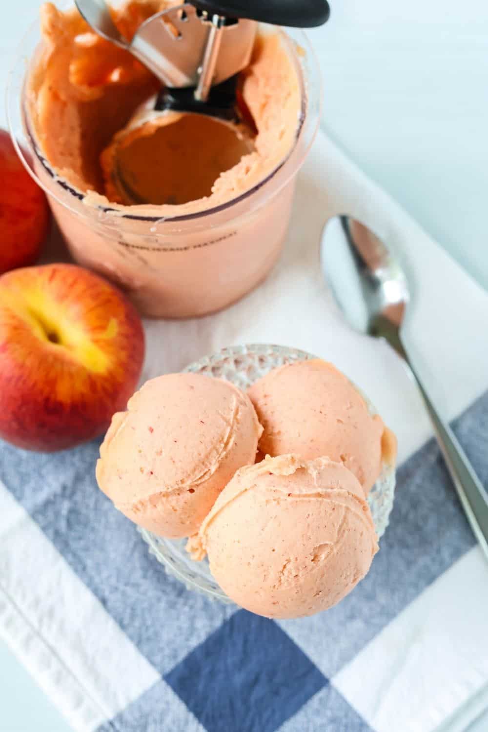 overhead view of a glass dish with three scoops of no-peel peach sorbet in it, with a Ninja Creami pint container in the background.