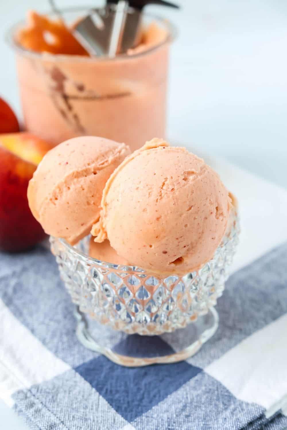 scoops of Ninja Creami fresh peach sorbet in a glass dish, with the pint container and peaches in the background.