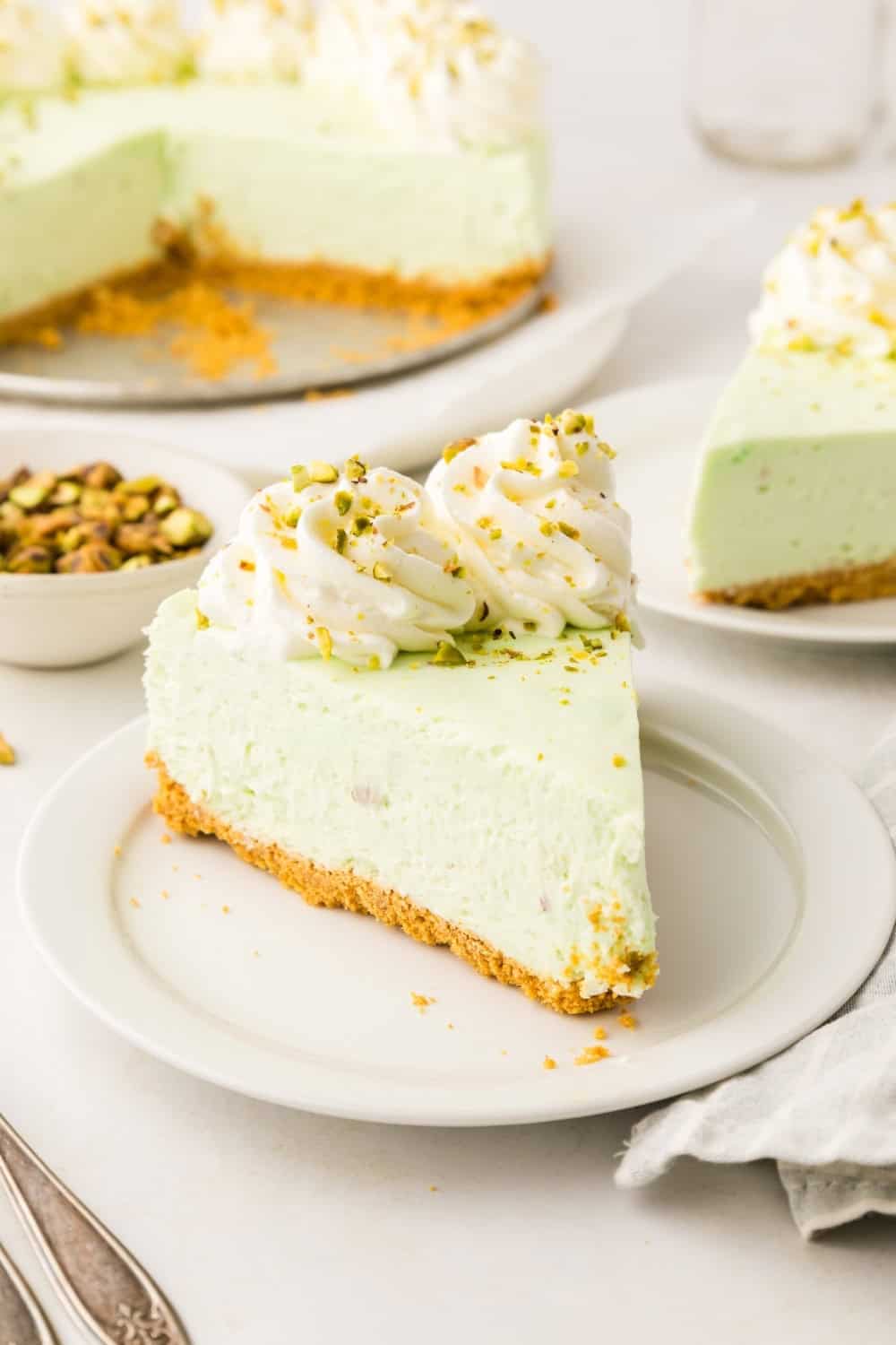 a slice of no bake pistachio cheesecake on a white plate, with the remaining cheesecake in the background.