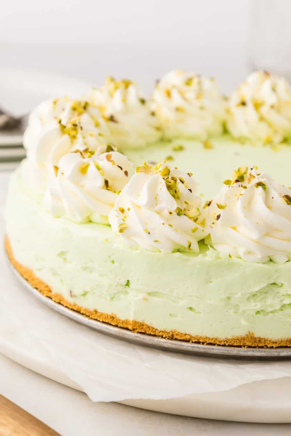 close-up side view of a pistachio cheesecake, garnished with swirls of whipped cream and chopped pistachio nuts.