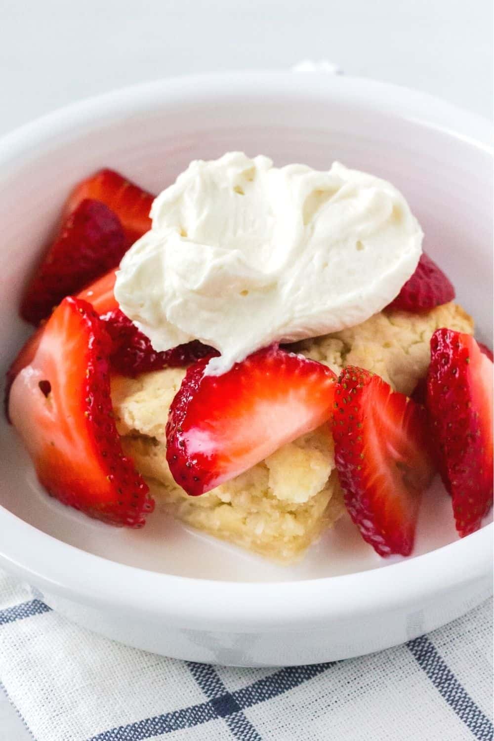 a white bowl contains homemade shortcake topped with strawberries, whipped cream, and a drizzle of milk.