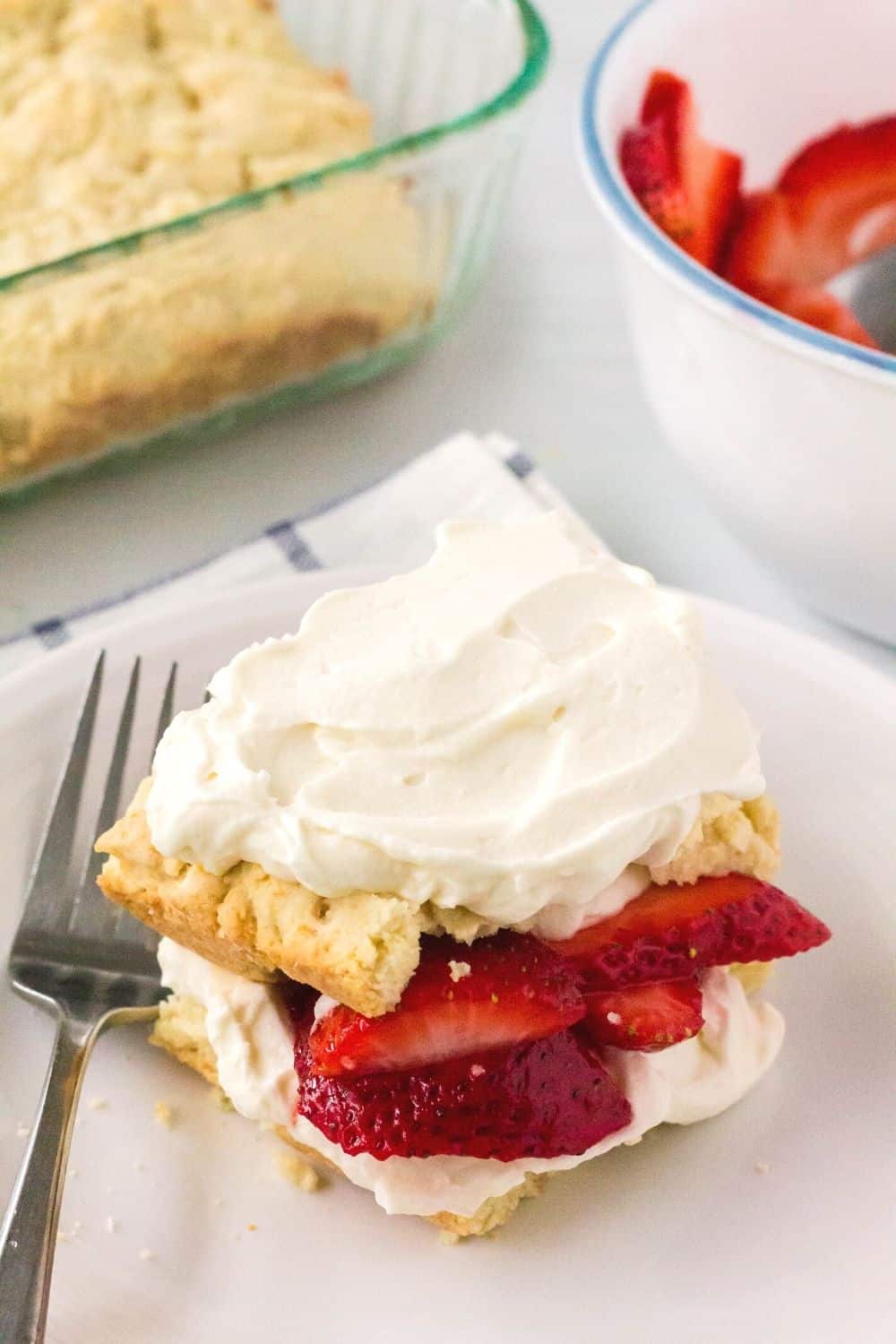 homemade shortcake topped with fresh strawberries and whipped cream. A bowl of strawberries and the remaining shortcake are in the background.