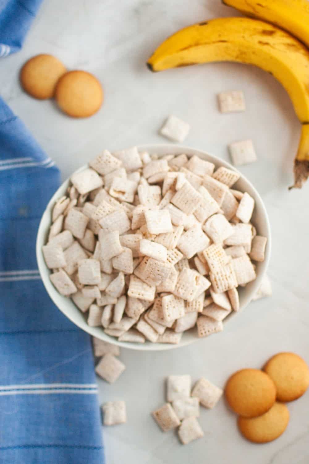 overhead view of banana pudding muddy buddies served in a white bowl, with vanilla wafers and bananas in the background.