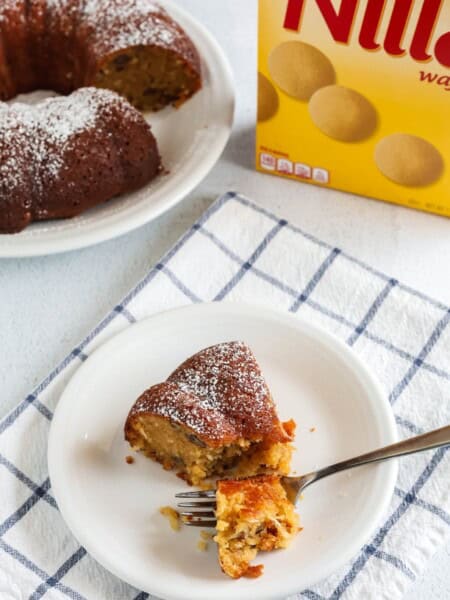a slice of vanilla wafer cake is served on a white plate with a fork; the remainder of the bundt cake and a box of Nilla Wafers is in the background.